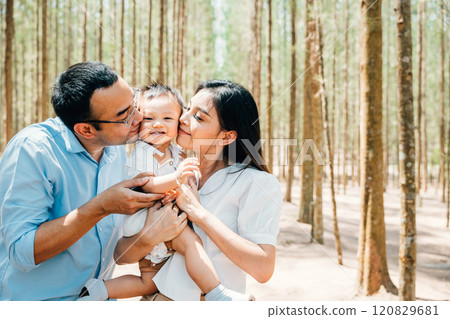 Asian family parents embracing in a lush green forest nature kissing child, happy father mother have picnic kiss his daughter together, Family holiday 120829681