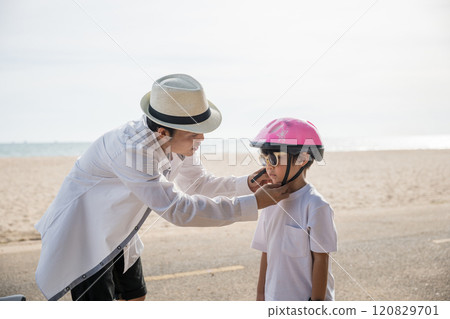 On a beach vacation a family experiences a heartwarming scene, the father in a safety helmet imparts the art of bicycle riding to his cheerful son creating an inspiring moment of learning and joy. On a beach vacation a family experiences a heartwarming scene, the father in a safety helmet imparts the art of bicycle riding to his cheerful son creating an inspiring moment of learning and joy. 120829701