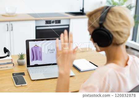 Happy Caucasian woman ordering clothes online using laptop while listening to music in headphones. Seated at wooden table with smartphone and notebook, she enjoys a relaxed shopping experience. 120832878