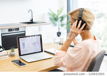 Happy Caucasian woman ordering cosmetics online on delivery website using laptop while listening to music in headphones. Bright modern kitchen setting adds freshness. Expression shows enjoyment. Happy Caucasian woman ordering cosmetics online on delivery website using laptop while listening to music in headphones. Bright modern kitchen setting adds freshness. Expression shows enjoyment. 120832903