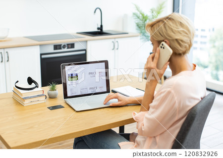 Caucasian woman using laptop and smartphone to order cosmetics online in modern kitchen. Concept of online shopping, convenience, and digital lifestyle. Emphasizing technology. Caucasian woman using laptop and smartphone to order cosmetics online in modern kitchen. Concept of online shopping, convenience, and digital lifestyle. Emphasizing technology. 120832926