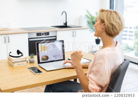 Caucasian woman in mid-30s cheerfully orders food online using laptop in modern kitchen. She appears happy and engaged in activity. Laptop screen displays food items with prices. Caucasian woman in mid-30s cheerfully orders food online using laptop in modern kitchen. She appears happy and engaged in activity. Laptop screen displays food items with prices. 120832953