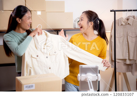 Two young asian woman live online clothing store while showing clothing with tablet and laptop. 120833107