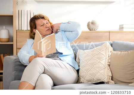 Elderly caucasian woman sitting on sofa reading book while pain neck with discomfort in living room. Elderly caucasian woman sitting on sofa reading book while pain neck with discomfort in living room. 120833710