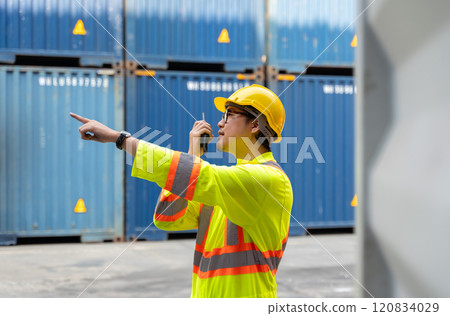 Asian man worker talking on walkie talkie for control loading containers box  while standing in front of blue containers at cargo shipping yard 120834029