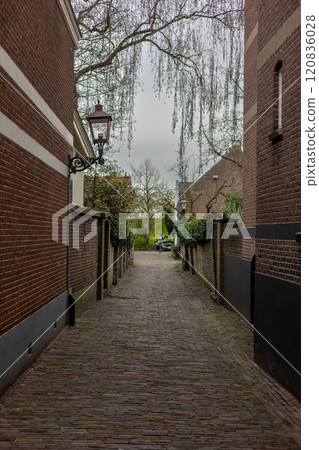 A narrow cobblestone alleyway flanked by brick buildings, with a vintage lantern on the left wall, trees in the background, and a cloudy sky overhead, creating a calm, historic atmosphere. A narrow cobblestone alleyway flanked by brick buildings, with a vintage lantern on the left wall, trees in the background, and a cloudy sky overhead, creating a calm, historic atmosphere. 120836028