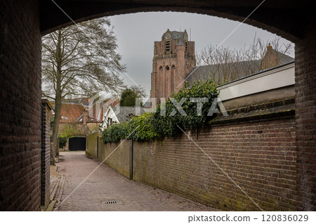 A cobblestone alley framed by brick walls leads to a view of a historic church tower. Ivy-covered walls and quaint houses add charm under an overcast sky, creating a peaceful, timeless atmosphere. A cobblestone alley framed by brick walls leads to a view of a historic church tower. Ivy-covered walls and quaint houses add charm under an overcast sky, creating a peaceful, timeless atmosphere. 120836029