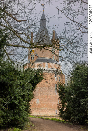 A historic brick castle with tall spire-like towers reflected in a calm moat, surrounded by leafless trees on a cloudy day, creating a serene and mysterious atmosphere. Castle Duurstede. 120836032