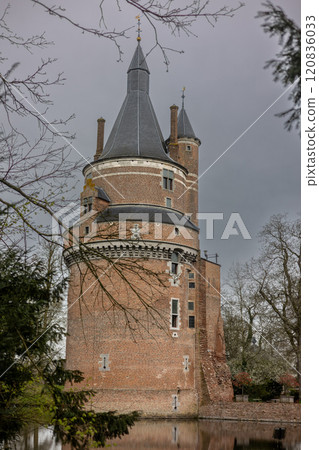 A historic brick castle with tall spire-like towers reflected in a calm moat, surrounded by leafless trees on a cloudy day, creating a serene and mysterious atmosphere. Castle Duurstede. A historic brick castle with tall spire-like towers reflected in a calm moat, surrounded by leafless trees on a cloudy day, creating a serene and mysterious atmosphere. Castle Duurstede. 120836033