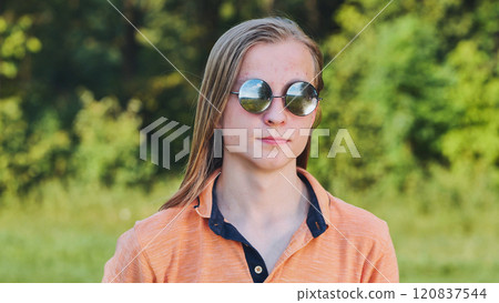 Portrait of a blonde teenager with long hair wearing sunglasses and posing in nature. 120837544