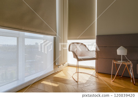 Modern automatic roller blinds and sun shades cover large windows, diffusing natural light into cozy room. Blackout and screen fabrics close-up. Space features stylish chair and minimalist side table. Modern automatic roller blinds and sun shades cover large windows, diffusing natural light into cozy room. Blackout and screen fabrics close-up. Space features stylish chair and minimalist side table. 120839420