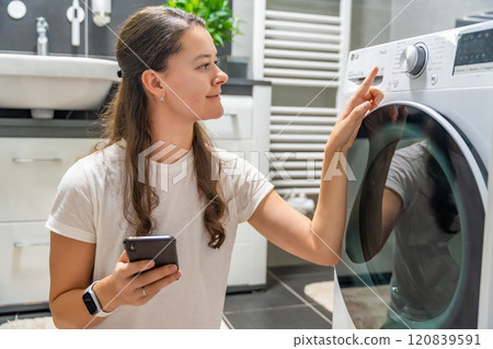 Young woman using mobile phone control via wi-fi and app for washing machine in home. Homesmart and new technology concept 120839591