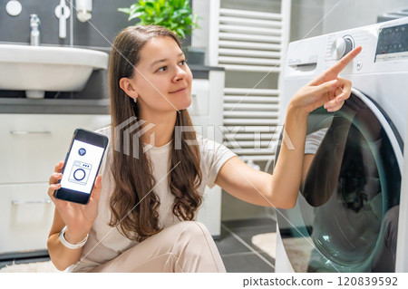 Young woman using mobile phone control via wi-fi and app for washing machine in home. Homesmart and new technology concept Young woman using mobile phone control via wi-fi and app for washing machine in home. Homesmart and new technology concept 120839592