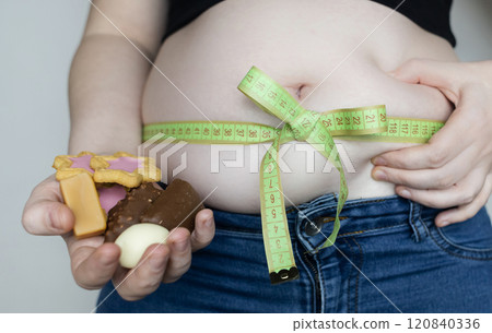 Obesity and overweight from overeating. Big belly tied with measuring tape as a gift. Body fat from candies and sweets Obesity and overweight from overeating. Big belly tied with measuring tape as a gift. Body fat from candies and sweets 120840336