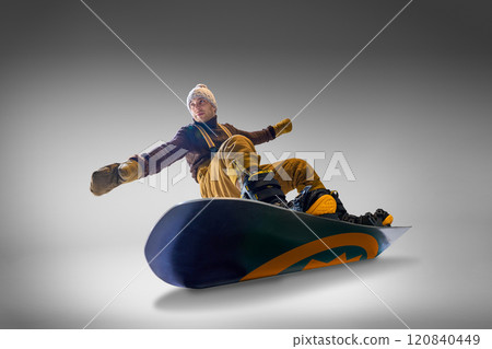 Snowboarder dressed in brown sweater and mustard pants leans back on his snowboard against gray background, showcasing balance and skill. 120840449