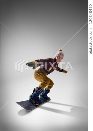 Snowboarder wearing cozy knitted sweater and gloves, mustard pants demonstrating stylish balance against grey studio background. 120840450