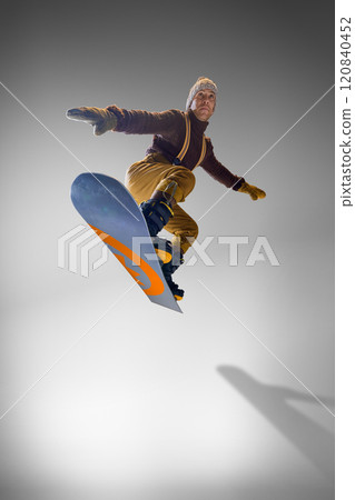 Snowboarder in brown sweater and mustard pants showcasing balance on his snowboard against grey studio background. 120840452
