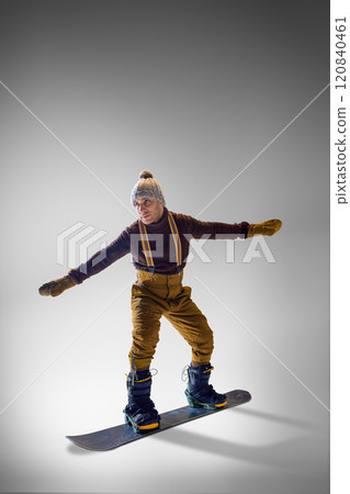 Athletic man, snowboarder in vintage-inspired winter wear balancing gracefully on his snowboard against grey studio background. Athletic man, snowboarder in vintage-inspired winter wear balancing gracefully on his snowboard against grey studio background. 120840461