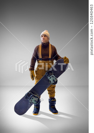 Snowboarder in brown knitwear and mustard pants holding his board confidently against grey studio background, ready for action. Snowboarder in brown knitwear and mustard pants holding his board confidently against grey studio background, ready for action. 120840463