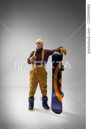 Man in a cozy sweater and knit hat posing with his snowboard against gray backdrop, showcasing winter style and adventure. Man in a cozy sweater and knit hat posing with his snowboard against gray backdrop, showcasing winter style and adventure. 120840464