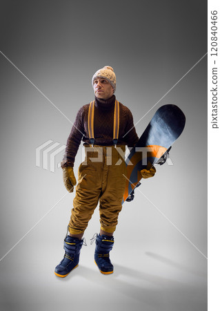 Snowboarder in brown sweater and mustard pants holding his board confidently against gray studio background. Snowboarder in brown sweater and mustard pants holding his board confidently against gray studio background. 120840466