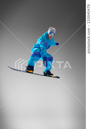 Snowboarder wearing bold outfit, jumps in mid-air against grey studio background, representing essence of seasonal adventures and extreme sports. 120840476