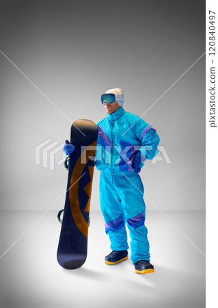 Young man, athlete dressed in blue retro-inspired suit posing with snowboard against grey studio background. Ideal for extreme sports themes. 120840497