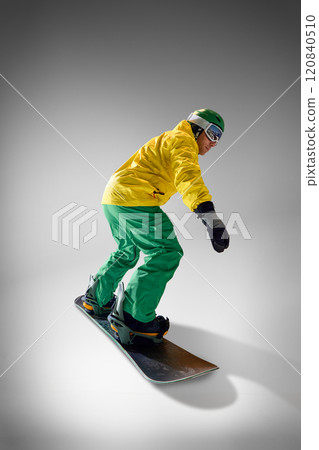 Side view, full length photo of athletic young man elegantly slopes on snowboard in motion, dressed vibrant clothes against grey studio background. Side view, full length photo of athletic young man elegantly slopes on snowboard in motion, dressed vibrant clothes against grey studio background. 120840510