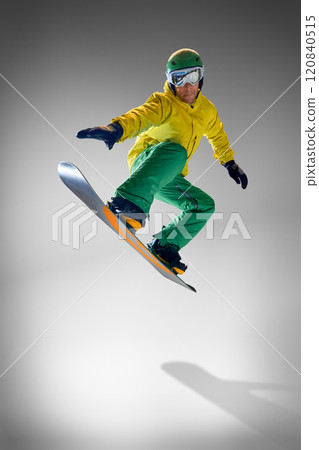 Young athletic man dressed modern, bright yellow and green snowboarding clothes performing tricks against grey studio background. 120840515