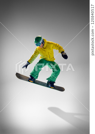 Young athletic man dressed vibrant yellow and green winter gear, performing tricks on snowboard against grey studio background. 120840517