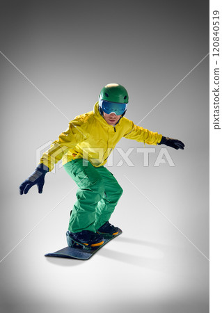 Snowboarder, young athletic man, in yellow jacket and green pants executing trick in mid-air against grey studio background. 120840519