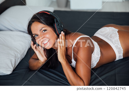Tanned woman in lingerie listening to music in headphones on bed in bedroom Tanned woman in lingerie listening to music in headphones on bed in bedroom 120842745