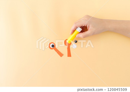 a hand holding a yellow glue stick, applying glue to an orange paper cutout. part of a craft project, creating a character or object with eyes 120845509