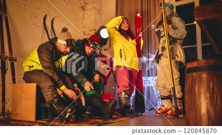 Group prepares for skiing indoors, tightening their boots and checking their gear, with vibrant outfits and festive setup of lights and decorations. 120845870