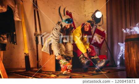 Two women laugh and prepare for skiing indoors, adjusting their boots while surrounded by festive lighting, vintage skis, and cozy winter decor. Two women laugh and prepare for skiing indoors, adjusting their boots while surrounded by festive lighting, vintage skis, and cozy winter decor. 120845871