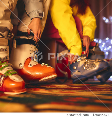 Close-up of ski boots being tightened as two people prepare for winter fun, their colorful outfits and holiday ambiance adding warmth to scene. Close-up of ski boots being tightened as two people prepare for winter fun, their colorful outfits and holiday ambiance adding warmth to scene. 120845872