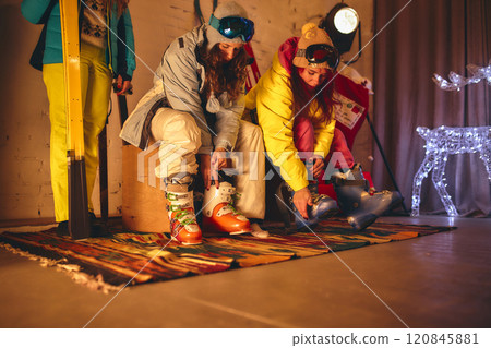 Two friends in colorful ski gear sit on bench indoors, fastening their ski boots, surrounded by cozy festive atmosphere with winter decorations. 120845881