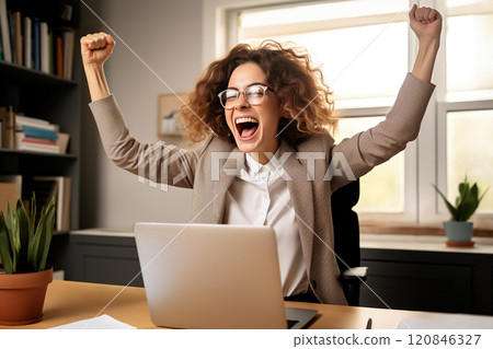 Excited and joyful young businesswoman at her desk with a laptop, celebrating a successful 120846327