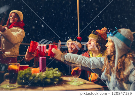 Friends bundled in winter gear toast with drinks outdoors as snow falls, sharing smiles and festive warmth in cozy holiday outdoor setting. 120847189