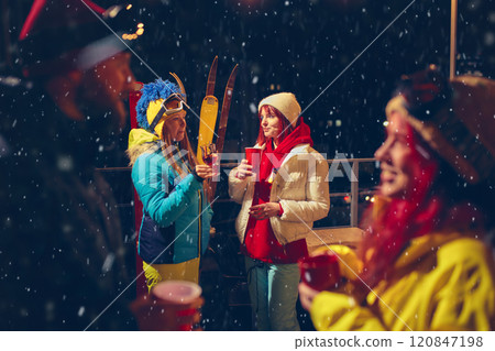 Cheerful group toasts outdoors on snowy evening, wearing colorful ski gear and sharing warm drinks, surrounded by festive, joyful vibe. 120847198
