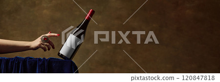 Mockup showing red wine bottle with blank label, being gracefully balanced by hand adorned with decorative red-stoned ring, against rich brown textured background. 120847818