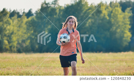 Young man is walking through a field on a sunny summer day, holding a soccer ball after a casual game with friends. 120848230