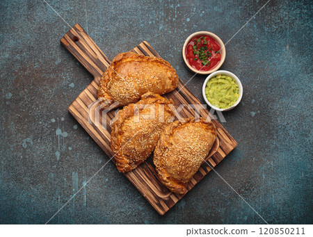 Delicious baked empanadas with sesame seeds served on wooden board with sauces, traditional Spanish savoury pastry Delicious baked empanadas with sesame seeds served on wooden board with sauces, traditional Spanish savoury pastry 120850211