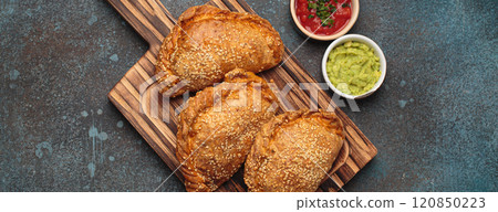 Delicious baked empanadas with sesame seeds served on wooden board with sauces, traditional Spanish savoury pastry Delicious baked empanadas with sesame seeds served on wooden board with sauces, traditional Spanish savoury pastry 120850223