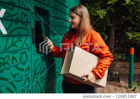 Young woman picks up parcel from automatic post office machine, standing with phone near the self-service mail terminal. Parcel delivery machine. 120851987
