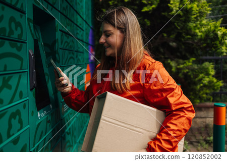 Young woman picks up parcel from automatic post office machine, standing with phone near the self-service mail terminal. Parcel delivery machine. 120852002