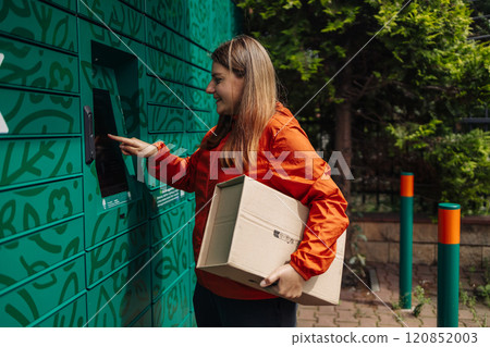 Young woman picks up parcel from automatic post office machine, standing with phone near the self-service mail terminal. Parcel delivery machine. 120852003