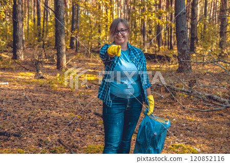 A female volunteer smiles while standing in the forest with a garbage bag in her hand and points her finger at the camera. The woman urges that you too can help save nature from garbage. 120852116