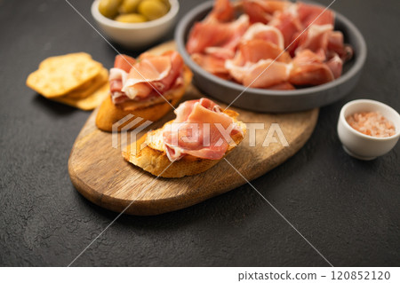 Italian sandwiches bruschetta with olives, bran bread, cherry tomatoes, prosciutto on wooden cutting board, dark black background surface Italian sandwiches bruschetta with olives, bran bread, cherry tomatoes, prosciutto on wooden cutting board, dark black background surface 120852120