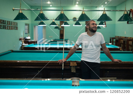 A happy handsome man stands in the billiard room, leaning on the table and chatting with friends. The man is having a blast having fun with his friends and playing billiards. A happy handsome man stands in the billiard room, leaning on the table and chatting with friends. The man is having a blast having fun with his friends and playing billiards. 120852200